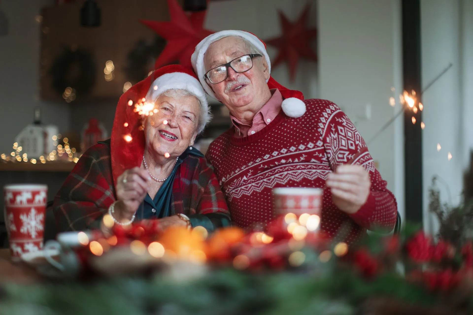 A joyful senior couple wearing Santa hats and holding sparklers while celebrating Christmas in a warmly decorated home.