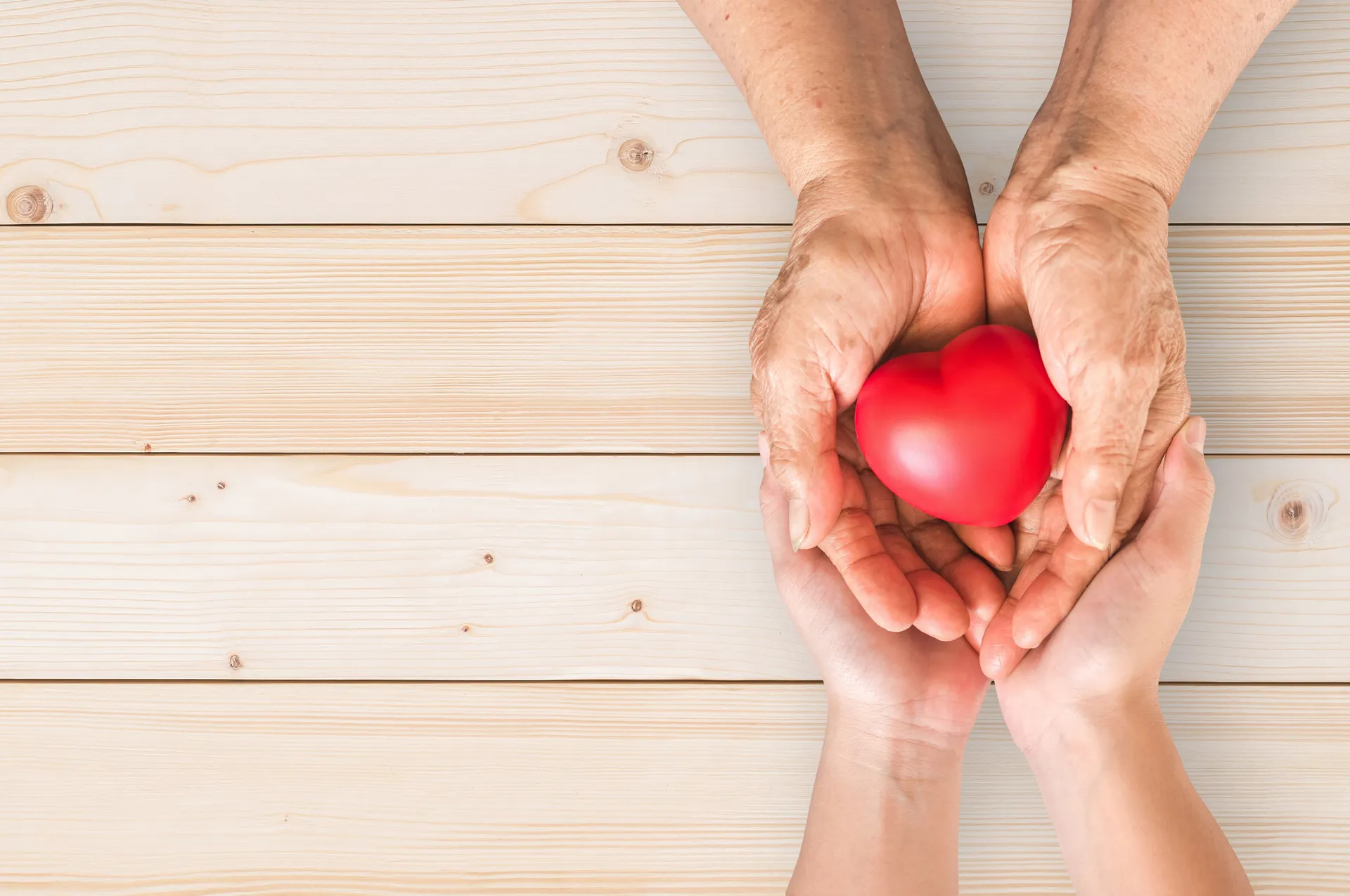Elderly hands and younger hands holding a red heart together, representing care, compassion, and health support.