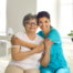 A caregiver sitting closely with an older woman as they smile and hug, showing companionship and emotional support.