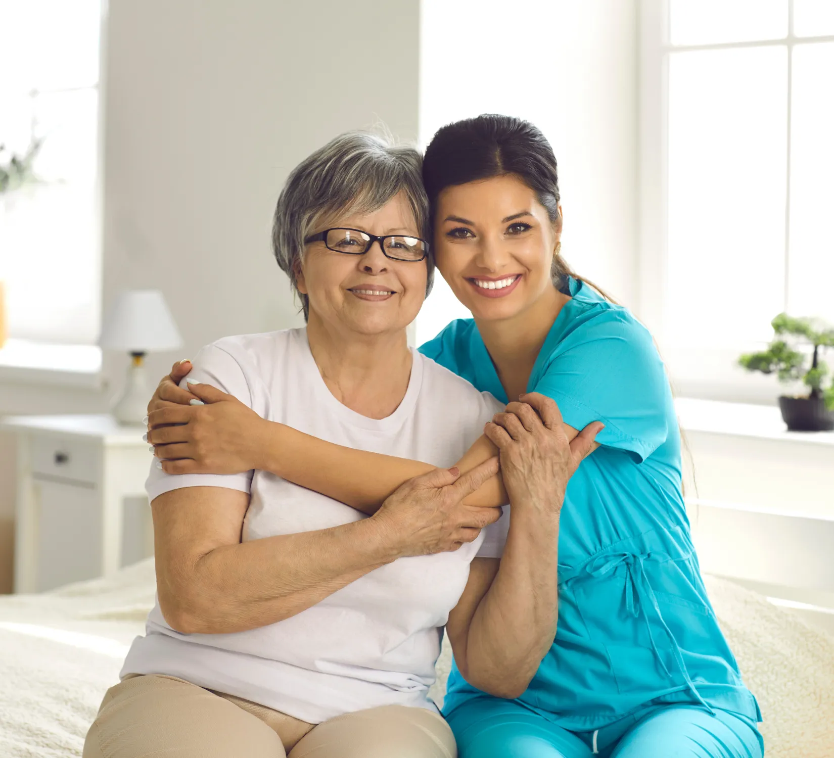 A caregiver sitting closely with an older woman as they smile and hug, showing companionship and emotional support.