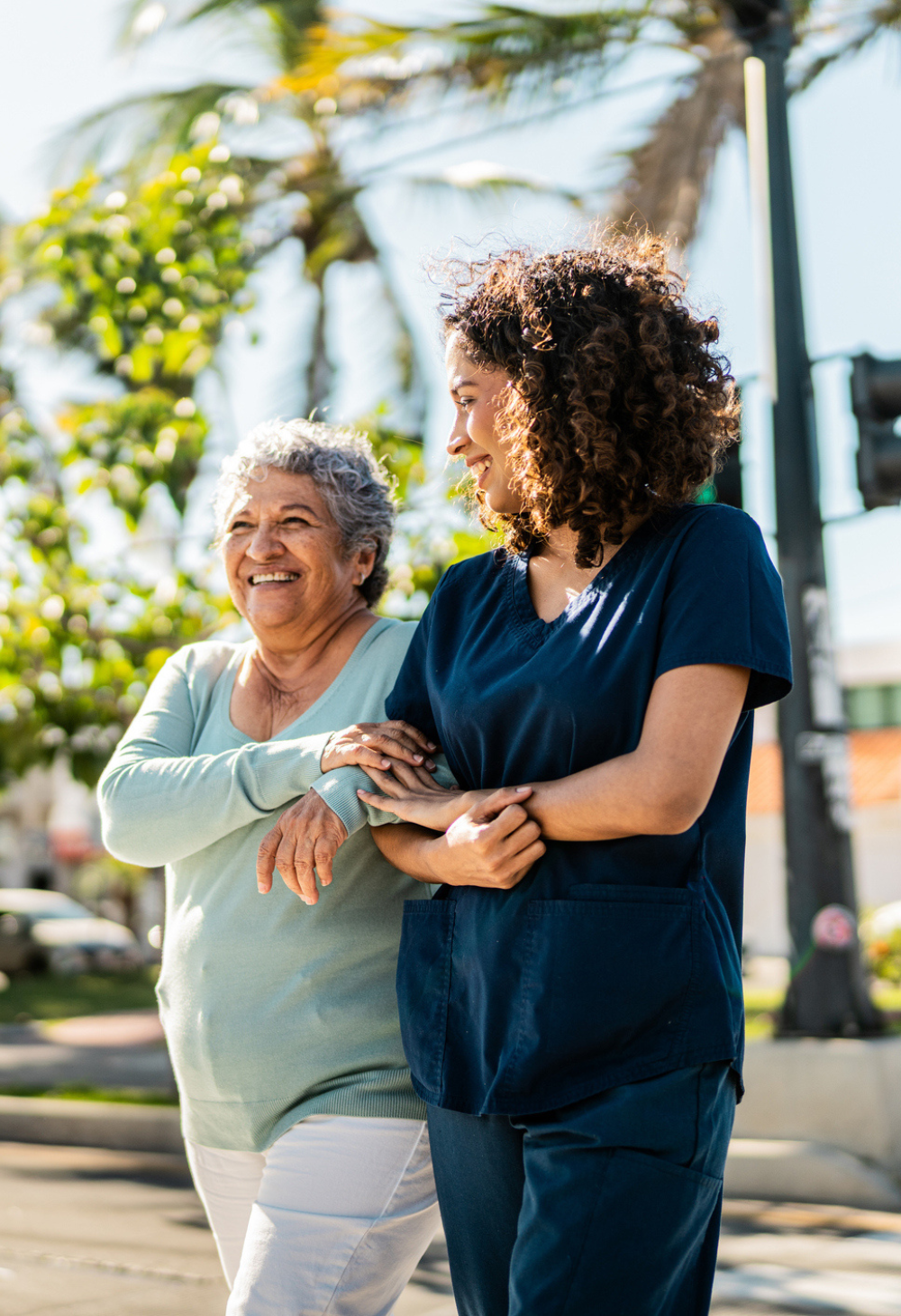 A caregiver walking arm in arm with an older woman outdoors on a sunny day, providing companionship and support during a neighborhood stroll.