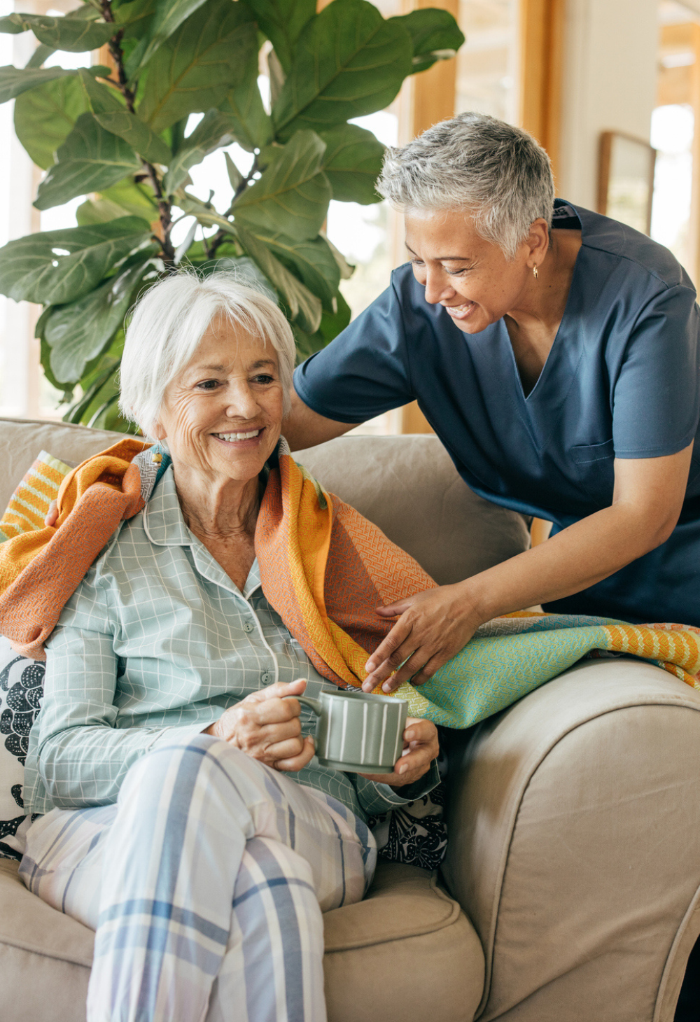 A caregiver gently placing a warm blanket over a smiling older woman who is sitting on a couch with a cup of tea, providing comforting in home support.