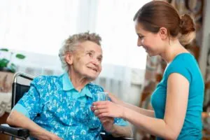 A caregiver handing a glass of water to an older woman in a wheelchair, providing hydration support and compassionate in home care.