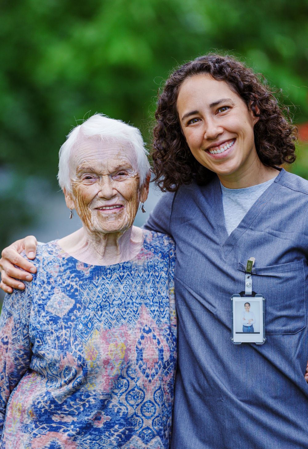 A caregiver in scrubs posing with a senior woman in an outdoor setting.