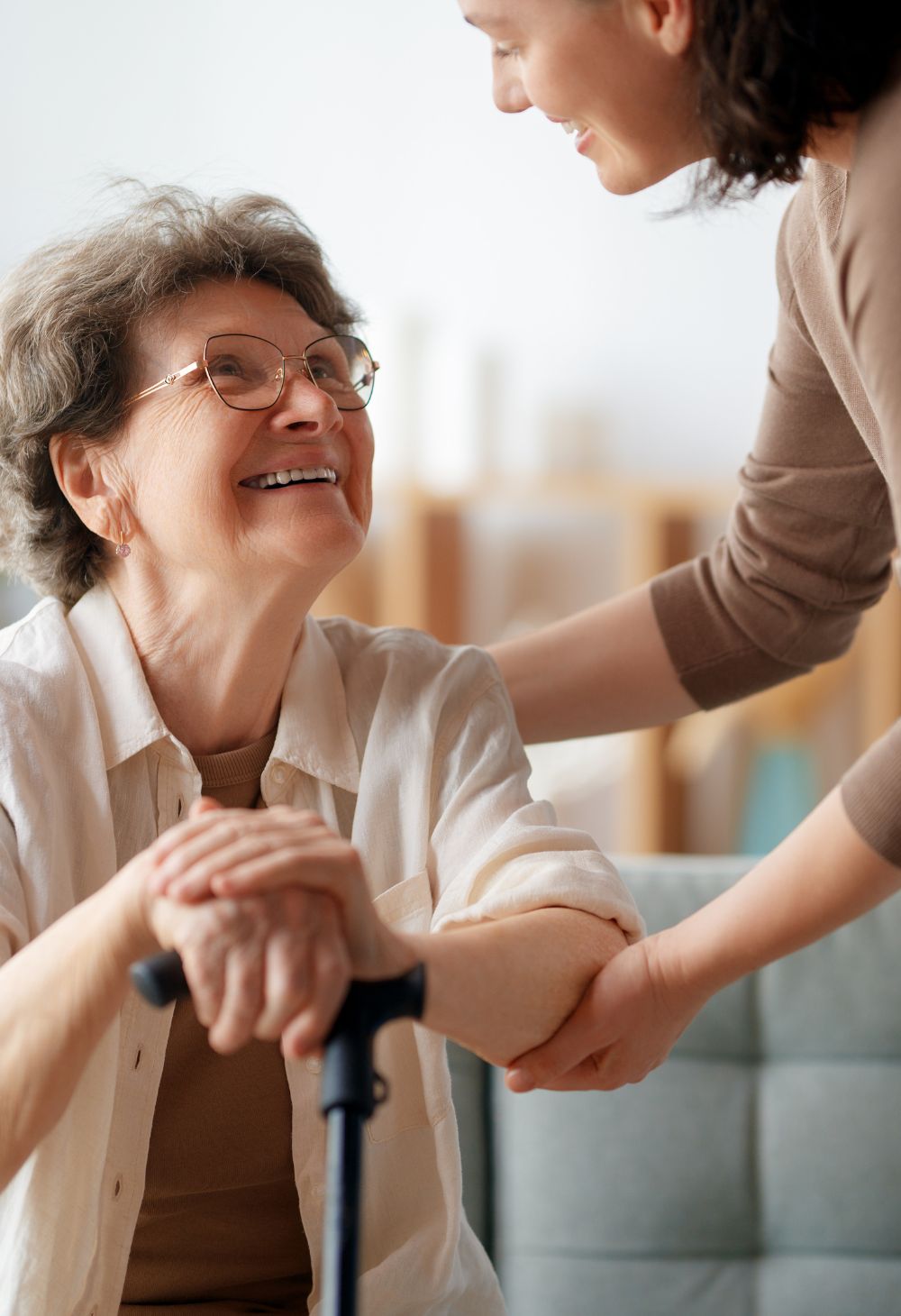 Caregiver offering support to an elderly woman holding a cane, smiling together in a warm home setting