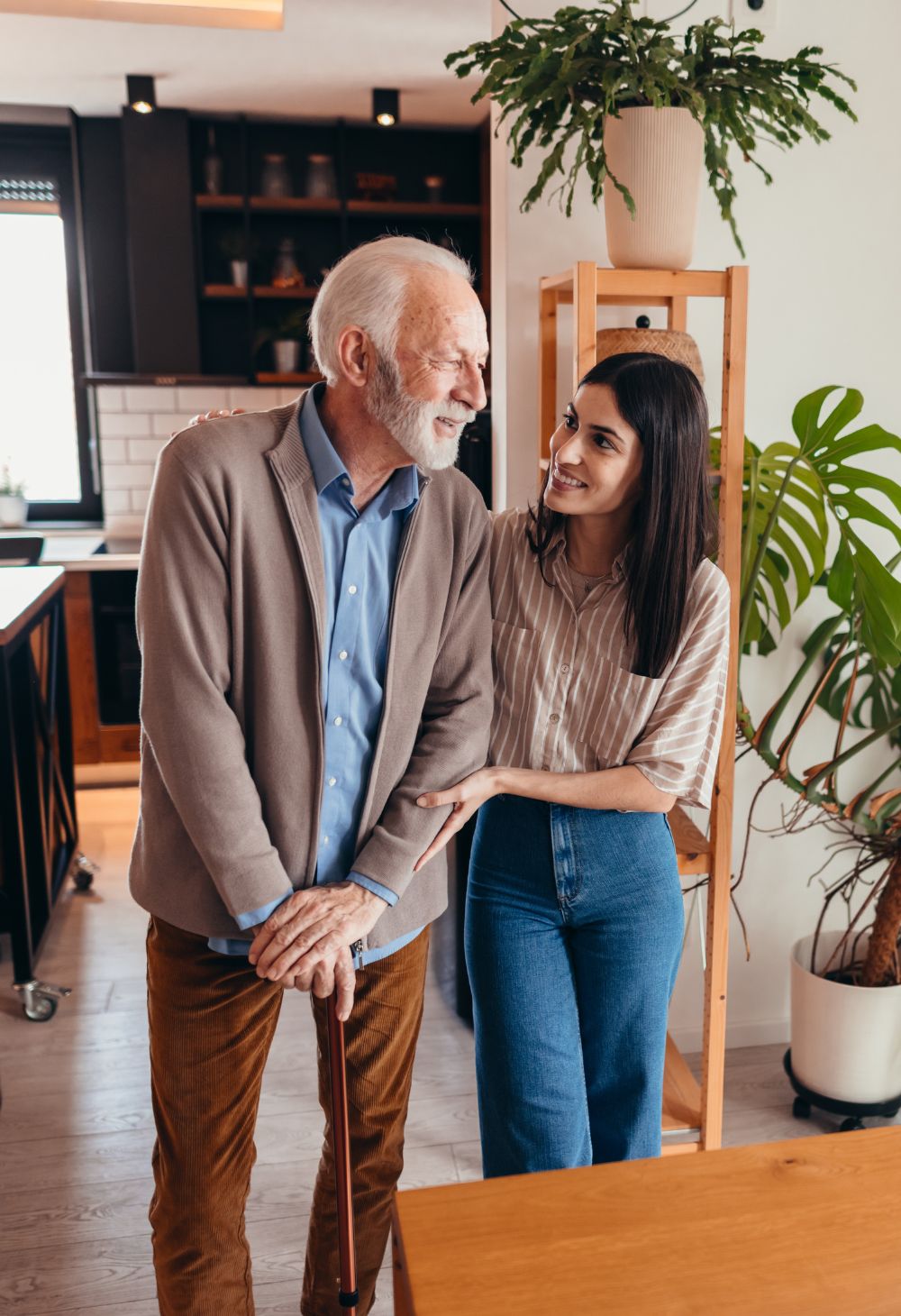 A caregiver gently holding the arm of an older man walking with a cane, providing steady support during an outdoor walk.
