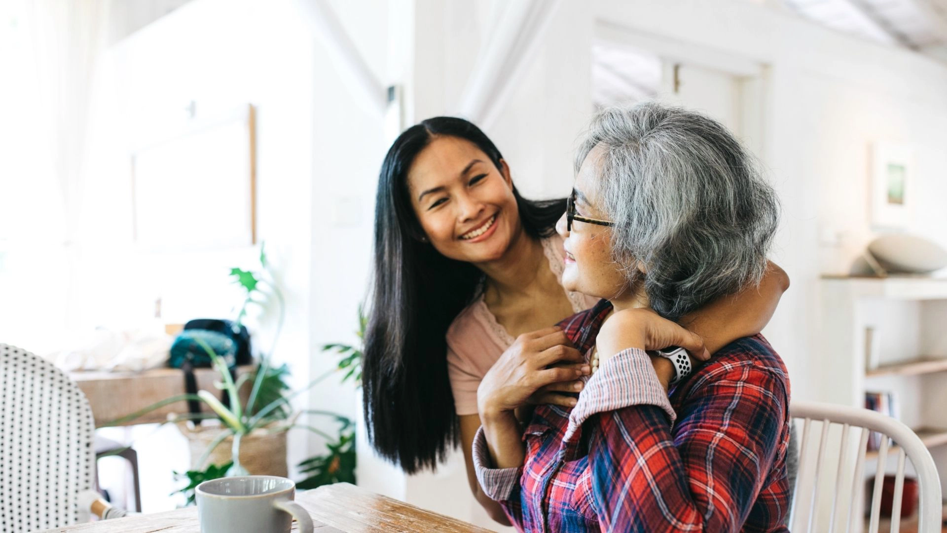 A caregiver standing beside a senior woman at a dining table, engaging in a warm and friendly conversation inside the home.