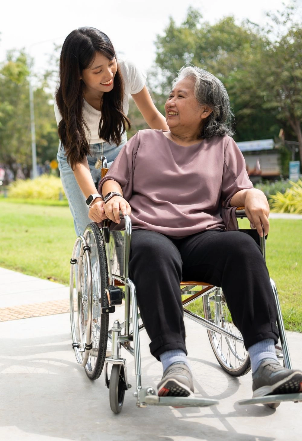 A young female caregiver pushing a smiling senior woman in a wheelchair along an outdoor path, providing compassionate support and companionship.
