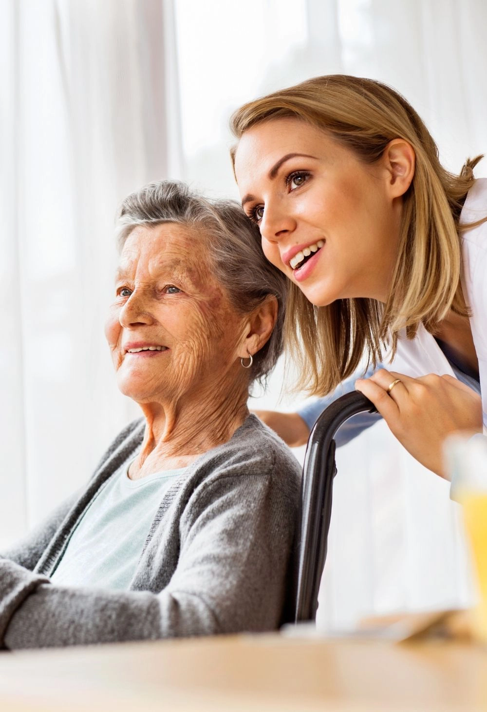 A smiling caregiver leaning beside an older woman seated indoors, offering emotional support and companionship.