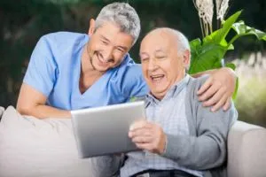 A caregiver sitting beside an older man while they look at a tablet together and laugh, demonstrating companionship and engaging activities for seniors.