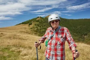 An older man wearing a helmet and using trekking poles while hiking through a sunny, grassy landscape, enjoying an active outdoor lifestyle.