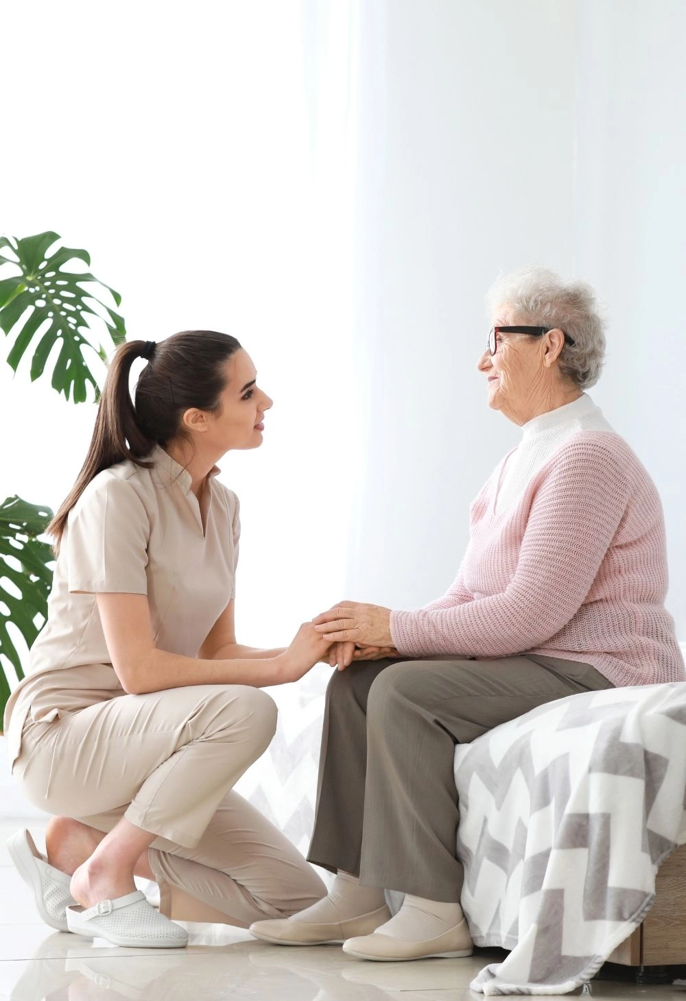 Caregiver Comforting Senior Woman at Home A caregiver kneeling beside an elderly woman sitting on the bed while holding her hands and offering emotional support.