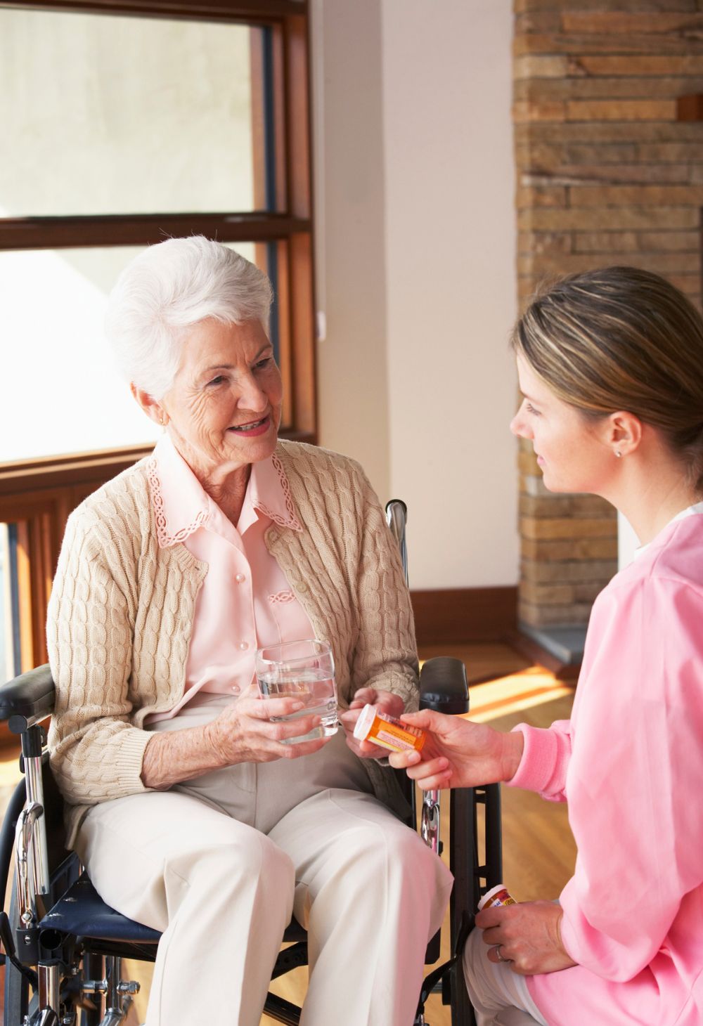 Caregiver assisting an elderly woman in a wheelchair with medication and a glass of water at home