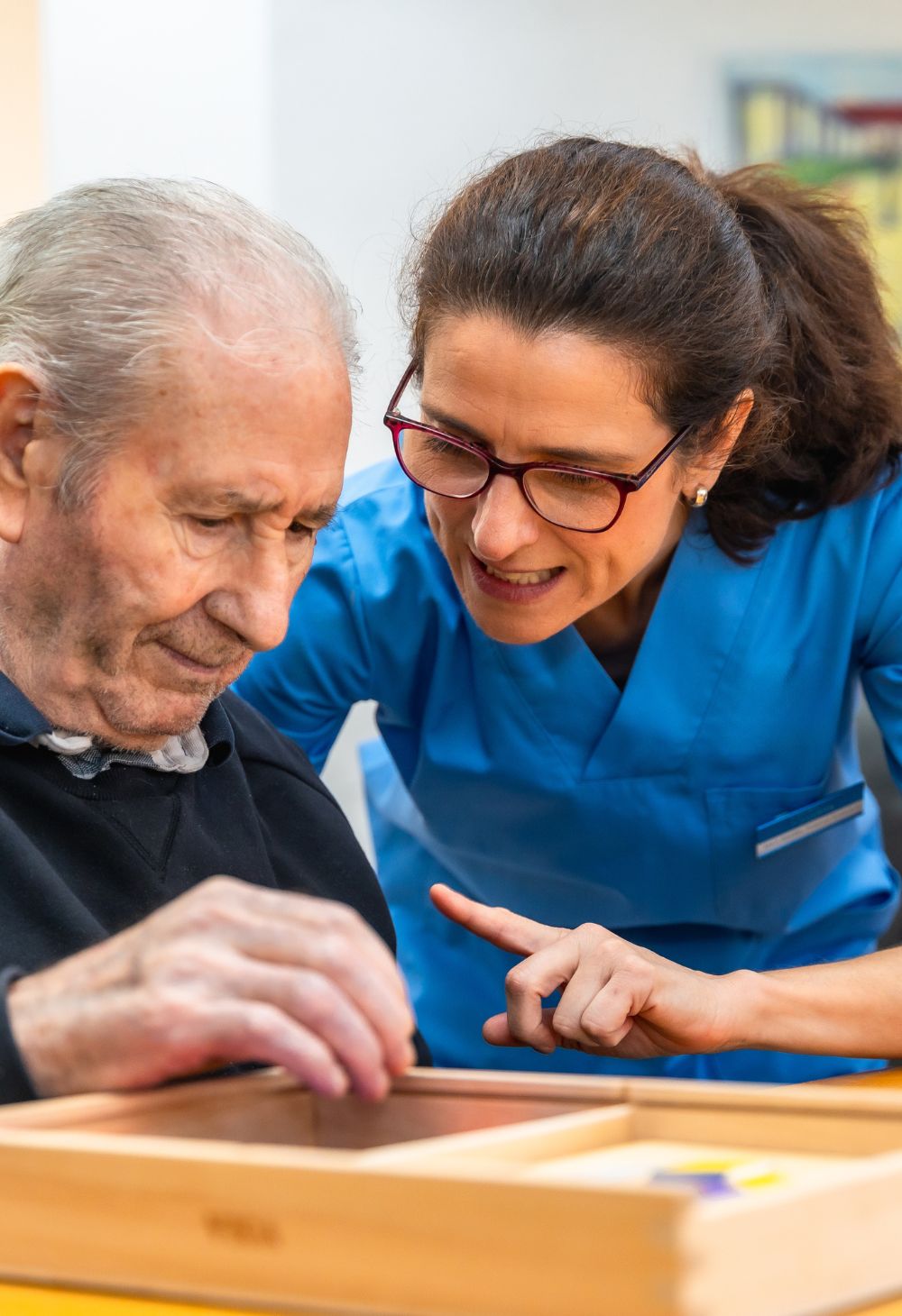 A caregiver in blue scrubs assisting an elderly man with a cognitive activity, guiding him with a gentle and supportive gesture.