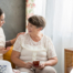 Smiling caregiver with her hand on an older woman’s shoulder as they share tea on the couch, showing home care vs assisted living