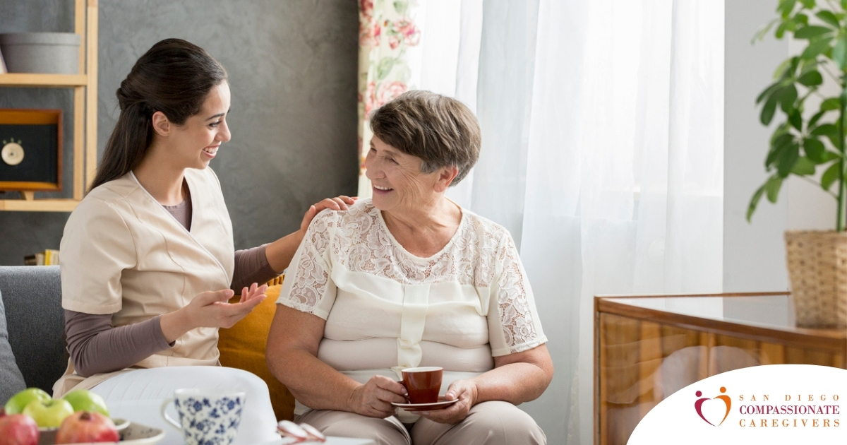 Smiling caregiver with her hand on an older woman’s shoulder as they share tea on the couch, showing home care vs assisted living